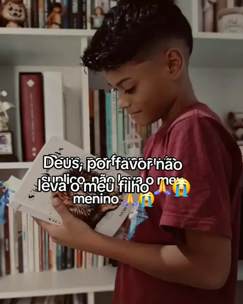 A young boy with short hair is sitting and reading a book, he is facing the camera and is holding the book. The background consists of a bookshelf filled with books. The boy wears a maroon shirt