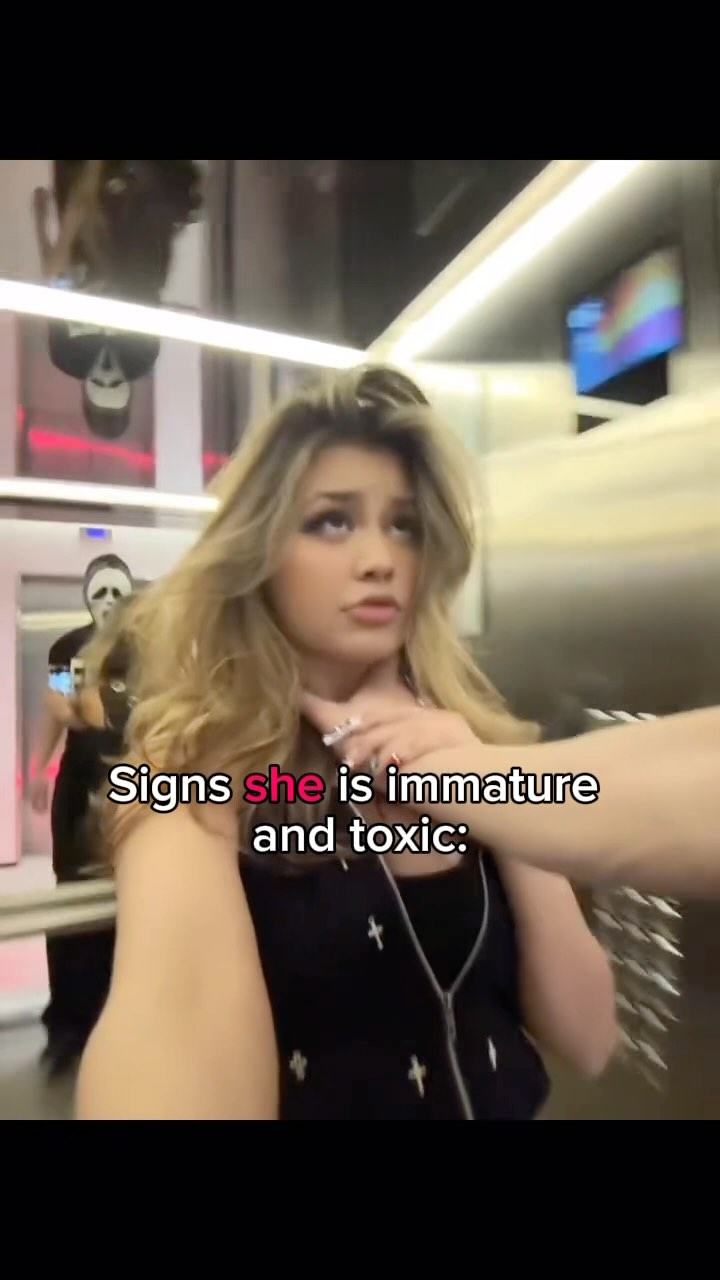 Selfie-shot in a shiny interior, with a young woman as the central element, looking towards the camera with a concerned expression. In the background, a person is partially visible, wearing a mask, adding an unclear element to the context. The lighting is bright, contributing to a modern, somewhat glossy feel, enhanced by the metallic surfaces behind.