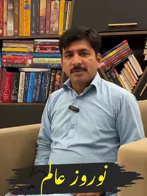 A man in a light blue shirt is seated in front of a bookshelf filled with books. The man has a mustache and is wearing a lavalier microphone. Overlaid on the bottom of the image is yellow text in an Urdu script.
