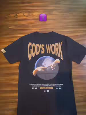 A black T-shirt is displayed on a wooden surface. The shirt has a design on the back featuring the words 'GOD'S WORK' in a gold, stylized font, arching above a circular image. Inside the circle, two hands are reaching toward each other, as in Michelangelo's 'The Creation of Adam'. Below the image is a scripture reference and a logo. Above the shirt is a purple cross symbol.