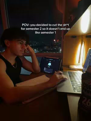 A young man is sitting at a desk, studying. He is looking at a laptop. The scene is set at night with city lights visible outside the window. There is a water bottle to the right.