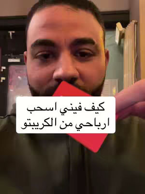 A selfie-style shot features a man against a slightly blurred background. The man is fair-skinned with dark hair, a neatly trimmed beard, and dark eyes. He's holding a bright red object in front of his face, partially obscuring his mouth, with only his eyes visible above it. The background includes indistinct elements and warm lighting suggesting an indoor setting. A white text box, with Arabic script, is superimposed onto the red object, enhancing focus on the text.