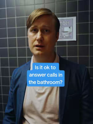 A man in a bathroom setting addresses the camera, wearing a blue blazer and a pink shirt. A text box overlaid on the image asks: "Is it ok to answer calls in the bathroom?" The background has black square tiles.