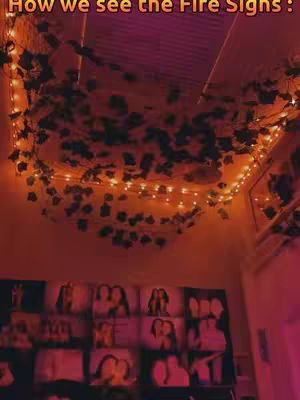 A low-angle shot of a room's ceiling and upper walls, primarily lit with warm, orange and pink hues. Strands of fairy lights are draped across the ceiling, interspersed with leafy vines. The lower portion of the image shows an assortment of framed pictures arranged on a wall.