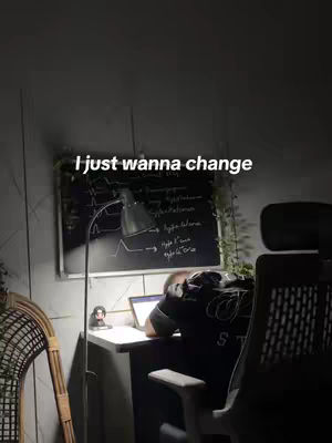 The image shows a dimly lit room with a desk and chair. A person is sitting at the desk with their face down and out of view. A chalkboard is behind them, with some writing on it. A lamp is lit, with the light shining on the desk. The overall impression is of someone studying or working late into the night.