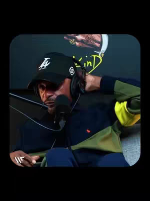 A man wearing a black LA baseball cap and headphones is speaking into a microphone. The background features a partial image of a person's hand with tattoos.
