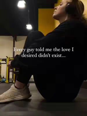 A woman is sitting in a gym setting with their head tilted upwards, eyes closed, and arms wrapped around their legs. White text overlayed on the image reads: 'Every guy told me the love I desired didn't exist...' The lighting seems to be from overhead fixtures, and the image has a slightly desaturated color palette.
