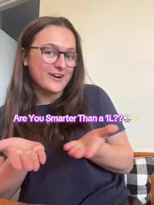 A woman with long brown hair and glasses smiles while gesturing with open hands. Text in a purple gradient overlay reads "Are You Smarter Than a 1L??⚖️✨". The background is a plain wall and a portion of a patterned cushion.