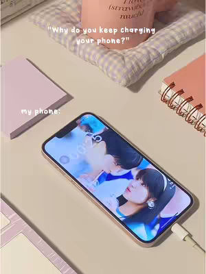 A top-down shot of a phone charging on a light-colored surface with various items arranged around it. The phone's screen displays a picture of two people, and the image is plugged into a charger. A pink notebook and a pink cup are in the background. The overall tone is soft and aesthetic.