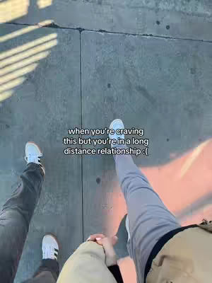 An overhead shot captures two people walking on a gray sidewalk. One person's legs are visible from the knees down, wearing light gray pants and white sneakers. The other person also has their legs in the frame, with dark jeans and white sneakers. They are holding hands. Text overlayed on the image reads: 'when you're craving this but you're in a long distance relationship :(' The lighting suggests a sunny day.