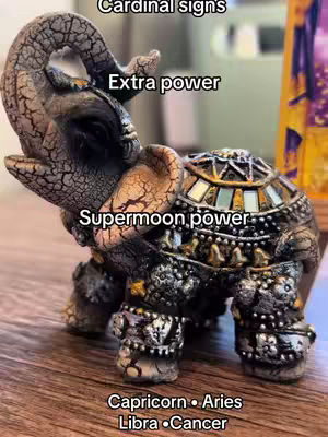 An ornate, brown and gold elephant figurine is the central focus, set on a wooden surface. Text overlays label the image, including "Cardinal signs", "Extra power", and "Supermoon power." Below the elephant, text labels "Capricorn • Aries Libra • Cancer". The background is blurred, suggesting an interior setting.