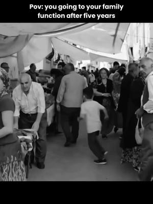 A black and white image depicts a crowded outdoor scene, likely a market or public gathering. People of various ages and attire are present, with some appearing to interact or move about. The composition is busy, filled with figures, stalls, and overhead coverings, creating a sense of a lively environment. The black and white tones add a sense of nostalgia or documentary-style realism.