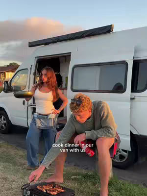 A couple is seen cooking dinner outdoors next to a white van on a sunny evening. The man is grilling food on a small portable grill, while the woman stands nearby. The van's door is open, and the background shows trees and a soft sunset.