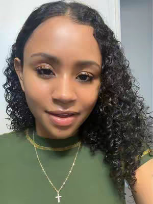A close-up shot of a woman with curly dark hair. She is wearing a green shirt and a gold necklace with a cross pendant. The background is a plain light color, and she is looking towards the camera with a slight smile.