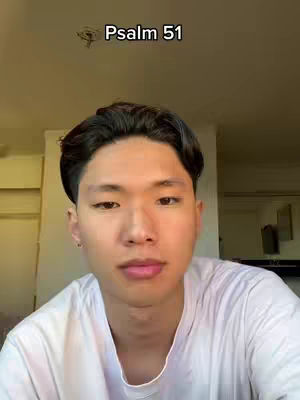 A young Asian man is centered in the frame, looking directly at the camera. The top of the frame shows text that says, 'Psalm 51'. The man is wearing a white t-shirt and the background is slightly blurred. The lighting seems soft, highlighting the man's face.