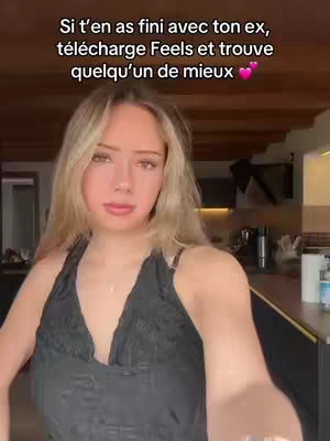 A young woman smiles and poses casually for the camera in a casual setting. She has long blonde hair and is wearing a black top against a warm interior background. Text overlays on the image promote a dating app.