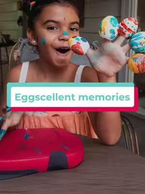 A young girl with a painted face and fingers, holding up her hand with painted Easter eggs on her fingertips. A red container sits in front of her.