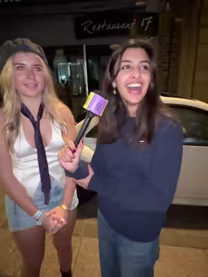 Two young women are seen outdoors, one holding a microphone and appearing to interview the other, who is smiling and looking at the camera. They are in front of a building at night, possibly restaurant.