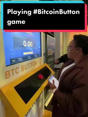 A woman in a brown coat stands right, holding a phone, in front a yellow kiosk with a large red button, a screen displaying a digital interface showing '0.00' and 'binance' logo. A black text overlay reads 'Playing #BitcoinButton game' in a white font, positioned above the woman. The background includes other blurred individuals and interior.
