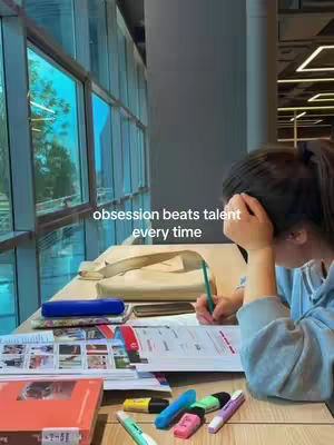 A study space with a large window overlooking a cityscape. A person is seated at a desk covered with books, papers, and colorful markers, writing with a pen while resting their head on their hand. The text overlay reads 'obsession beats talent every time'.