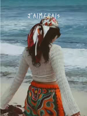 A woman stands on a beach, seen from the back, with the ocean in the background. She wears a white sweater and colorful patterned pants, with a headscarf. The sky is overcast.