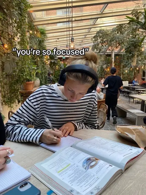 A woman is sitting at a wooden table, focused on writing in a notebook. An open textbook lies in front of her, while she wears headphones and is surrounded by a cafe setting with greenery and other people in the background. A text overlay reads: "you're so focused".