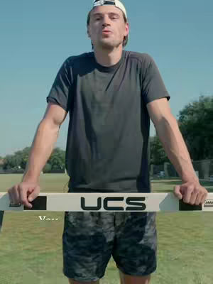A man stands in the foreground, holding a hurdle, with a field and blue sky in the background. The man is wearing a white cap, a dark t-shirt, and patterned shorts. The hurdle has the letters "UCS" on it. The overall composition is bright and sunny.