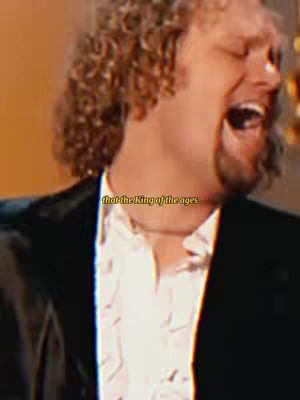 The image features a man with curly blond hair, seen from the chest up. He is smiling and singing with his mouth wide open. He wears a black blazer over a white, patterned shirt, and there is text overlayed on the image.