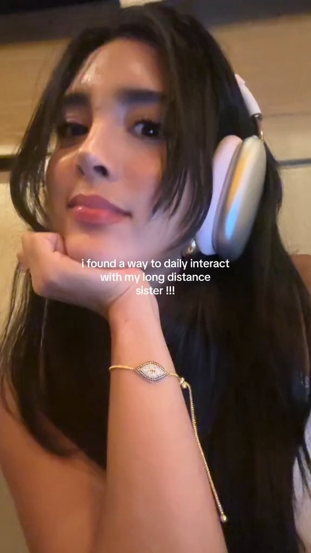 A selfie-style shot featuring a young woman with long dark hair and headphones. She's resting her head on her hand, looking at the camera with a slight smile. Text overlayed on the image says, 'I found a way to daily interact with my long distance sister !!!'.