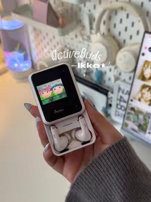 A hand holds a white flip-style case, showcasing white earbuds. The case's screen displays a cartoon image. The background features a pegboard with various items and text overlayed on the image.