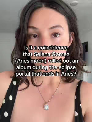 A woman with dark hair and a patterned top is centered in a selfie-style video. Text is overlaid on the image, asking if Selena Gomez releasing an album during an eclipse portal is a coincidence. The background is blurred, suggesting an indoor setting.