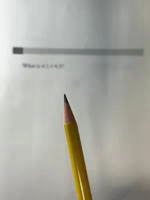 A close-up shot featuring a yellow pencil with its lead pointed towards the viewer. The pencil is positioned in the foreground, with a blurred background showing a sheet of paper and faint text, part of which reads, 'What is 1 + 1?'. The composition highlights the pencil's sharp tip against a simple backdrop.