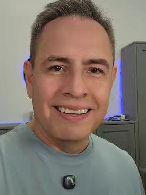 A man with short gray hair smiles at the camera. He wears a blue-gray shirt and a small black microphone is clipped to his collar. The background is a bright room with neutral-colored cabinets.