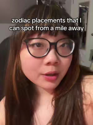 A person with long brown hair and bangs is centered in a selfie-style shot. They wear glasses and appear to be speaking. Overlaid text in the cover image reads 'zodiac placements that I can spot from a mile away.' The background is blurred, suggesting an indoor setting.