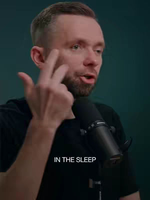 A man with short, light brown hair and a trimmed beard is speaking into a microphone. He is wearing a black t-shirt. The background is a dark teal color. He is pointing near his face with three fingers of his left hand. The text overlayed is a white color
