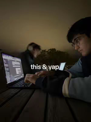 A low-angle shot at night shows two people studying at a wooden picnic table. A blurred figure with headphones sits to the left, while a person on the right, the primary focus, is wearing glasses, turned toward the camera, and using a laptop. The laptop screen is lit, displaying code or notes.