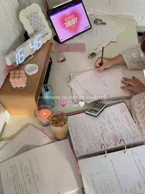 The image shows a well-lit workspace, likely a desk, filled with study materials. A laptop displays a heart-shaped graphic with the time 16:37. Papers, notebooks, a calculator, and a binder are scattered about, suggesting active studying. A hand is seen writing in a notebook, indicating someone is actively working. Various decorative items, such as candles, a digital clock, and a small container, add a personal touch to the study space, which features a drink and other small items.