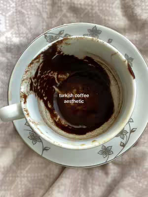 An overhead shot of a white cup and saucer set with decorative floral patterns. The cup contains dark coffee grounds in the center and the saucer is placed on top of a patterned cloth.