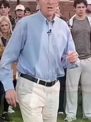 A man in a light blue button-down shirt and white pants is speaking in front of a crowd that is partially blurred. The man has a microphone attached to his collar.