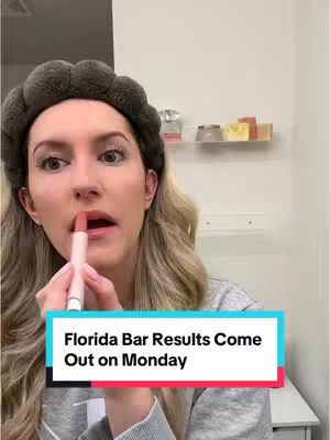 A woman is applying lipstick in front of a bathroom mirror. The image is well-lit, with a focus on the woman's face. The background includes a white wall, a shelf with various items, and countertop space. A text overlay at the bottom states, 'Florida Bar Results Come Out on Monday'.