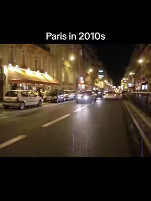 A nighttime street scene in Paris during the 2010s. The image is dominated by a wide road, with cars driving toward the viewer and parked along the sides. Buildings with illuminated windows and commercial establishments line the street, and streetlights cast a warm glow. The overall composition captures the essence of the city at night, with a focus on car traffic and architectural details.