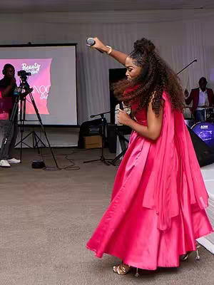 A woman in a bright pink dress with a microphone in her raised hand appears to be speaking, with her body angled towards the camera. Behind her is a screen with text, and a band setup is present. The lighting suggests an indoor event. The woman's hair is styled, adding to the formal vibe of the scene.