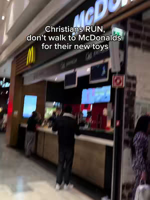 The image shows the interior of a McDonald's restaurant, with a focus on a counter and several people within the space. The McDonald's logo is visible, along with illuminated menu boards. A text overlay at the top states, 'Christians RUN, don't walk to McDonalds for their new toys.' The scene is captured from a low angle, emphasizing the depth of the interior.