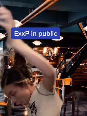 A woman with brown hair is in a public space, possibly a restaurant or bar, mid-action with her arm raised. She appears to be interacting spontaneously, with a blue rectangular text box overlaid on the image, reading "ExxP in public". The setting includes tables, chairs, and overhead lights, slightly blurred to emphasize the motion.