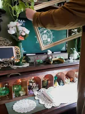 A cozy, maximalist setup is shown, featuring a monitor framed with an ornate gold frame held by a person. The scene includes a desk with various decorative items, a vase of flowers, and other whimsical objects.