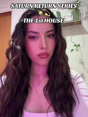 A selfie-style shot featuring a young woman with long, wavy brown hair, framed from the chest up. She gazes directly at the camera with a neutral expression. The background is softly lit with hints of interior decor, including a shelf with a plant. Text overlays at the top read "SATURN RETURN SERIES" and "THE 1st HOUSE".