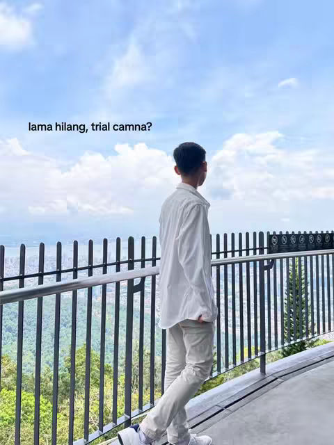 A person, viewed from the back and angled slightly to the side, stands on a walkway overlooking a hazy landscape. The individual is dressed in a white shirt and pants, with a scenic vista and cloudy sky filling the background. A black metal fence runs along the walkway's edge. A text overlay asking 'Lama hilang,trial camna?' is in the upper left.