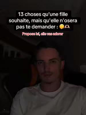 A man with short brown hair and a white t-shirt looks straight into the camera, with text on the screen above him. The image is framed up close, focusing on his face and upper body. The background is dark, possibly the inside of a room.