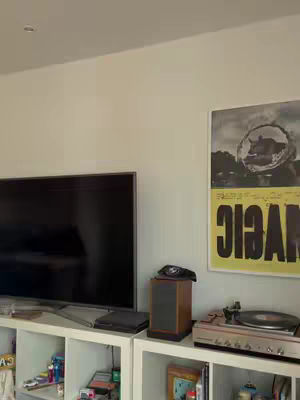 A room interior with a television, shelves containing items, a speaker, and a record player. A poster with the word 'MAGIC' is visible. The image is well-lit, with a neutral color palette.