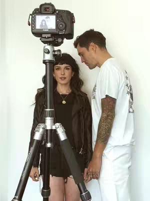 Two people are in front of a camera. A woman is standing in front of a tripod with the camera, and a man stands to her right. The woman has dark hair and is wearing a black outfit. The man has short hair, is wearing a white t-shirt, and has tattoos on his arm. They are near a white wall.